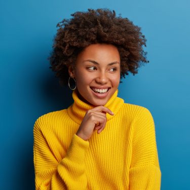 Positive curly young woman dressed in yellow comfortable sweater, holds chin, looks aside with dreamy expression, has interesting idea in mind, isolated over blue background. Ethnicity concept